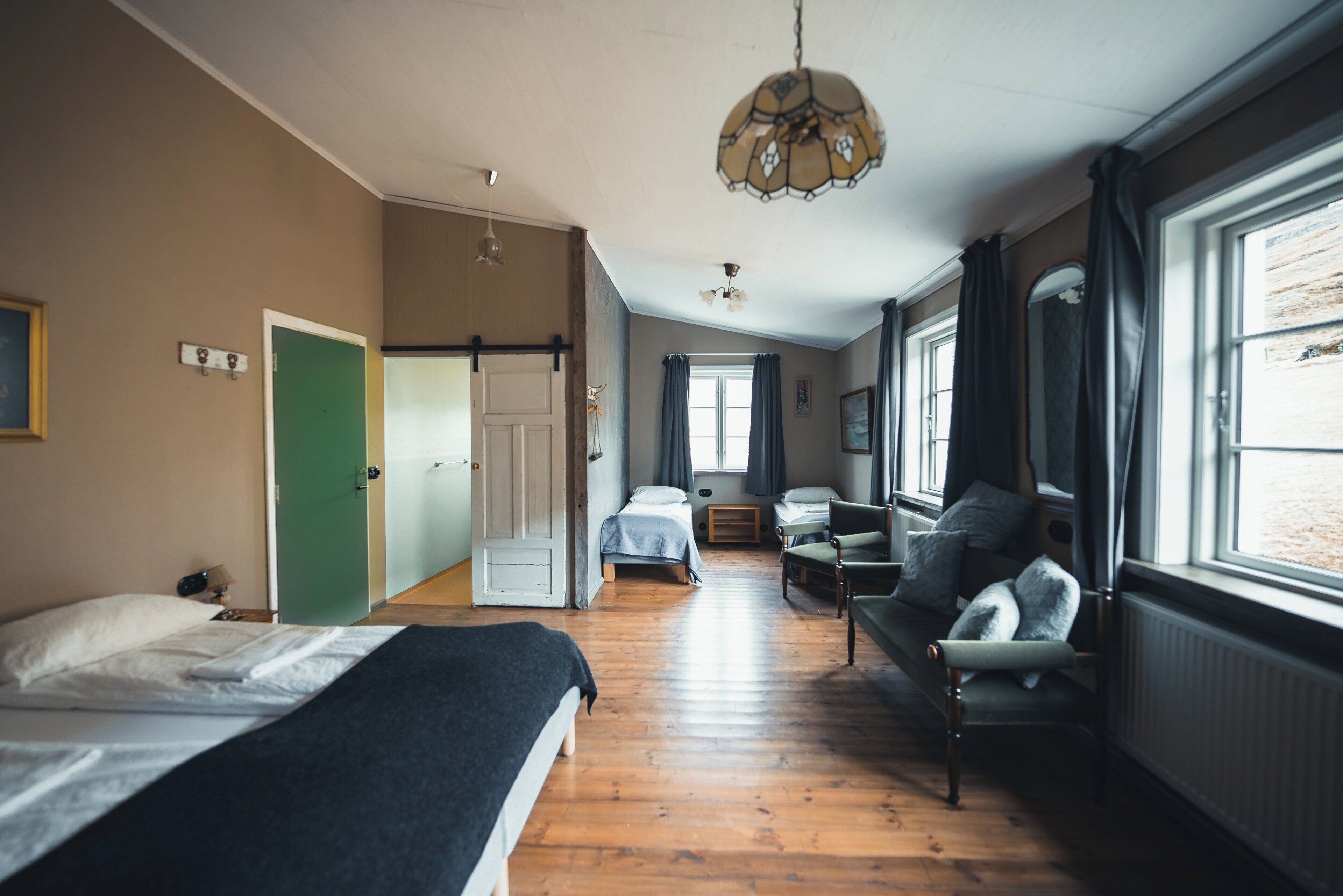 Una habitación con cama, sofá, sillón, ventana con cortinas y puerta.