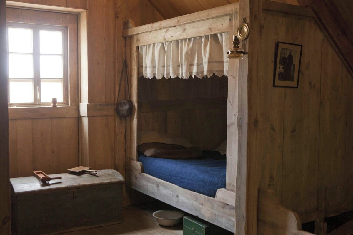 Une cabane en bois avec un lit superposé, une petite table avec des livres, une fenêtre et un tableau encadré au mur.