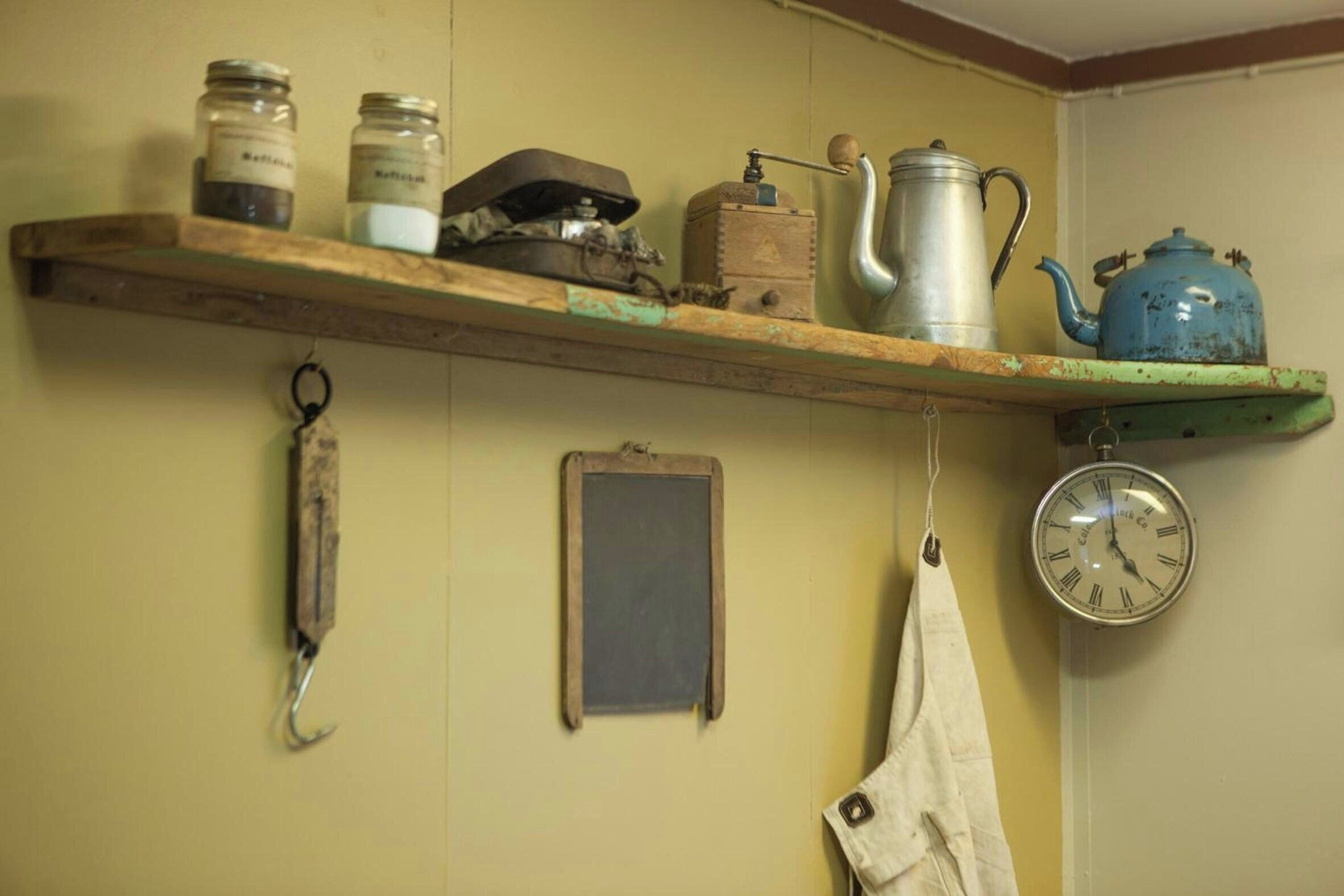 A kitchen shelf with jars, a kettle, a clock, and a hanging cloth.