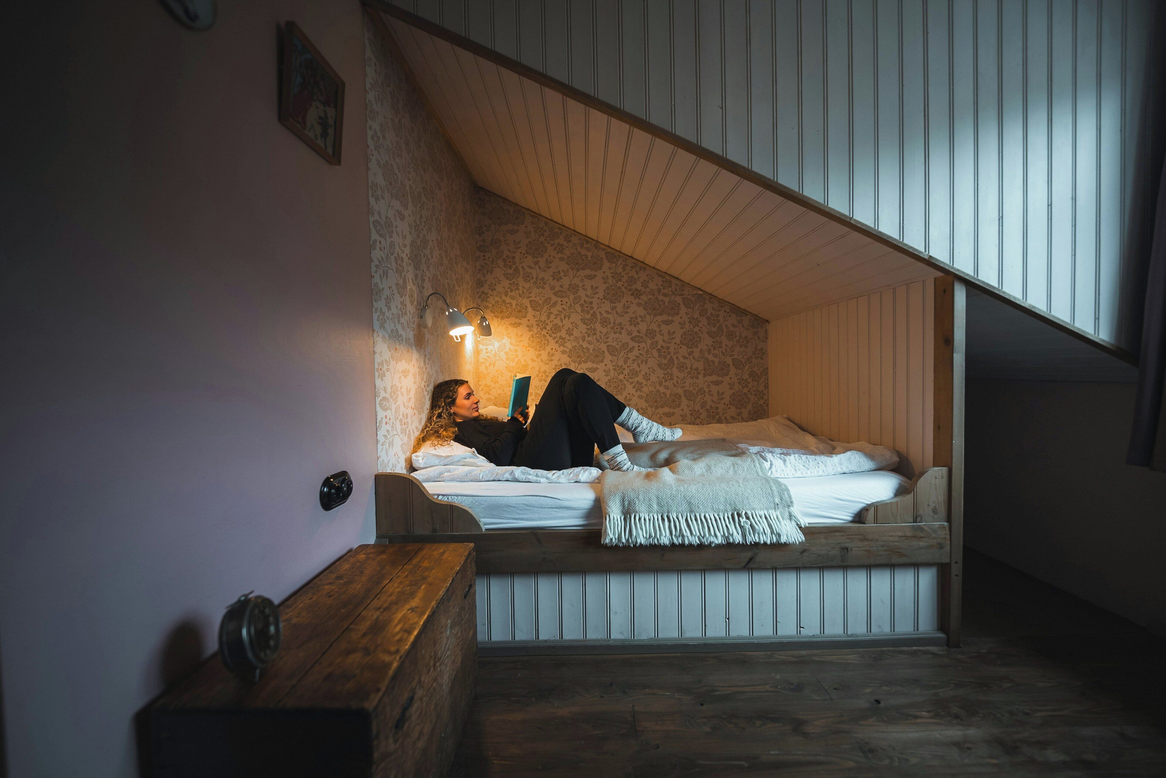 Un dormitorio acogedor con techo inclinado, una cama con edredón blanco, una lámpara de noche y una mesita de noche de madera.