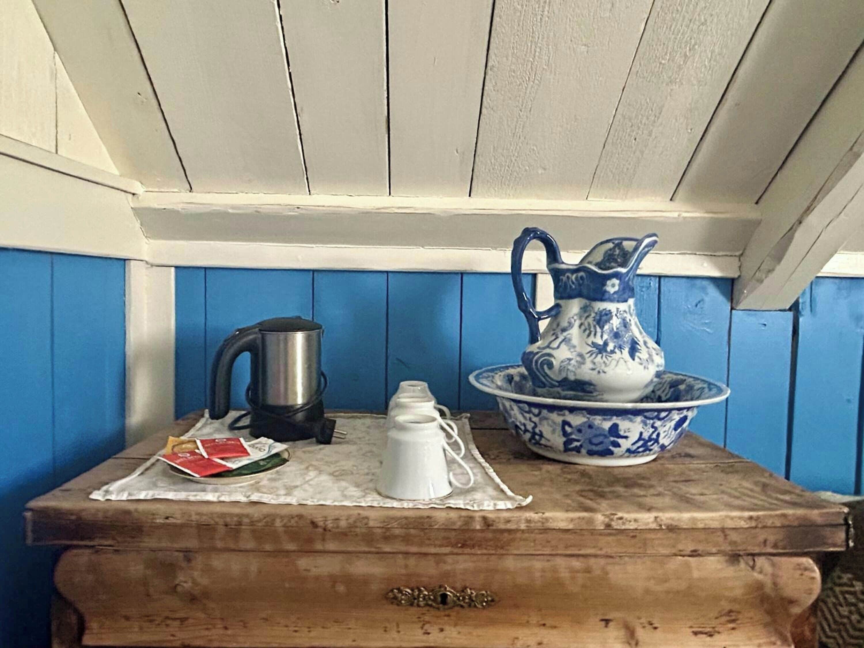 A wooden chest with a white cloth, a black kettle, a white pitcher, and a blue and white ceramic jug on top.