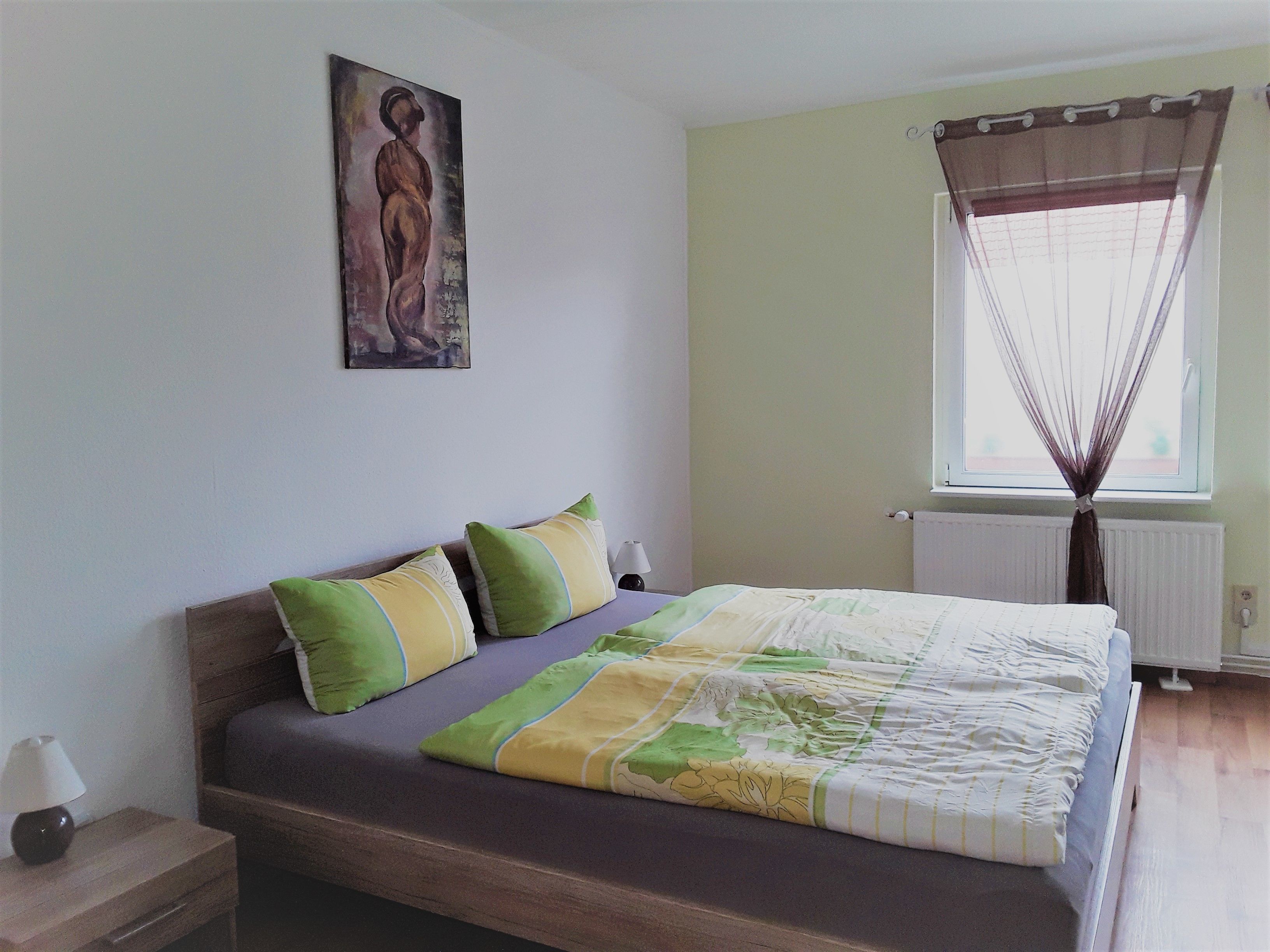 A bedroom with a bed, pillows, a nightstand, a painting, a window with curtains, and a radiator.