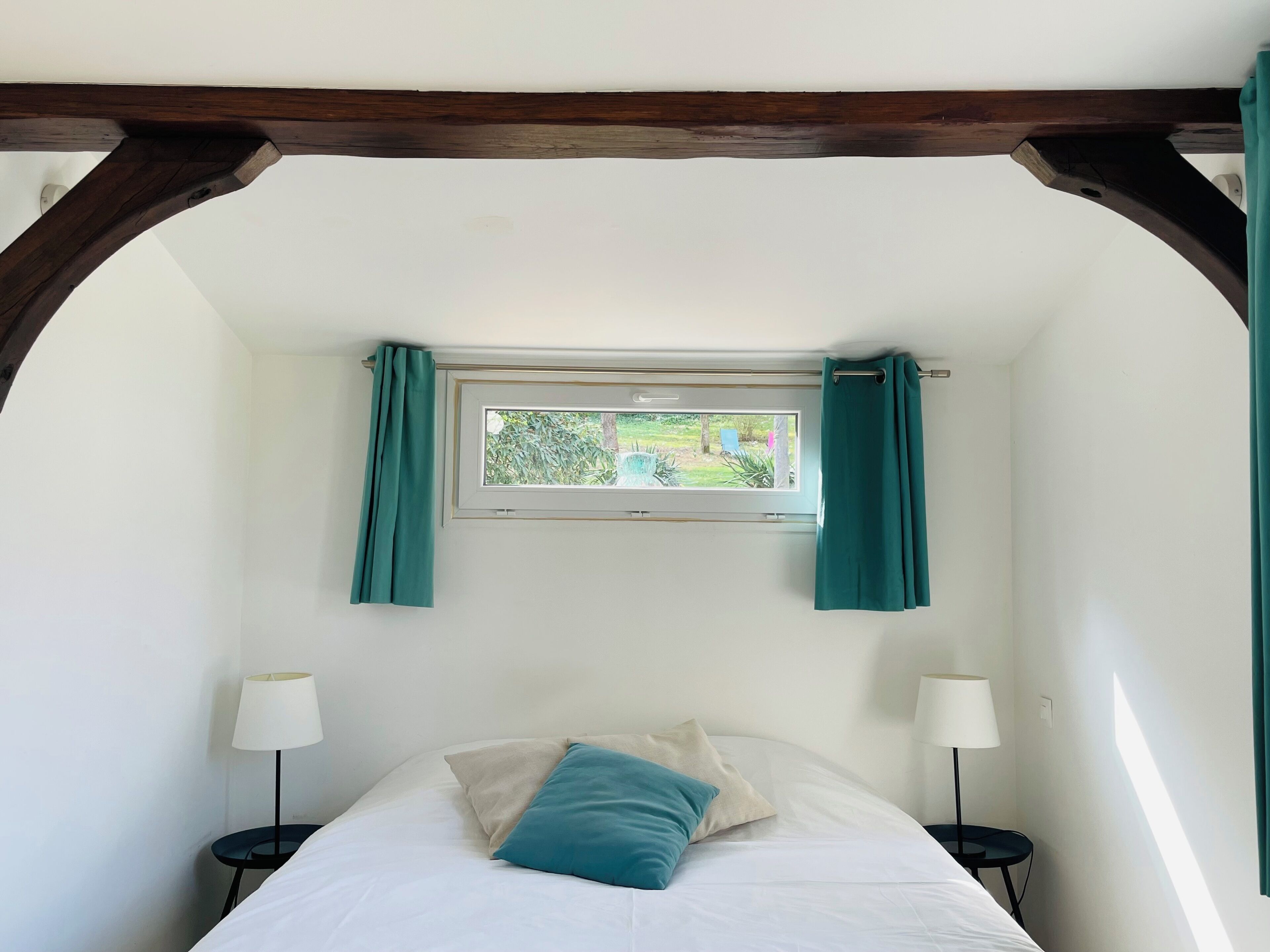 A bedroom with a bed, two bedside tables, a window with curtains, and a wooden ceiling beam.