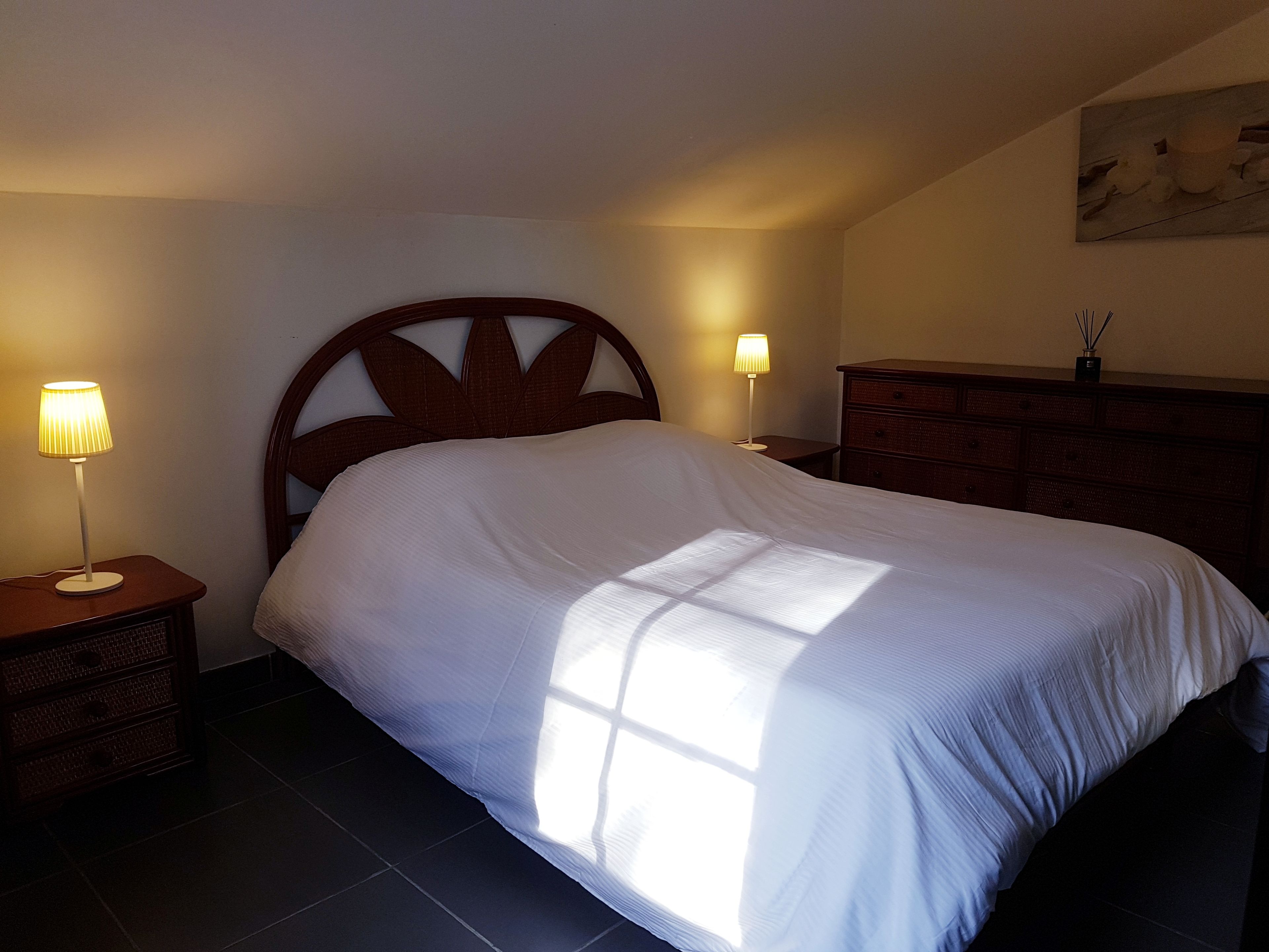 Un lit bien fait dans une chambre d’hôtel, avec une tête de lit en bois, deux lampes de chevet, une commode et un tableau accroché au mur.
