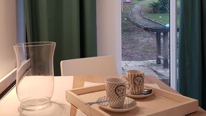 A table with a clear glass vase, two mugs, and a tray with a spoon. A window with green curtains reveals a garden view. - Ambition Zen Presles (Presles)
