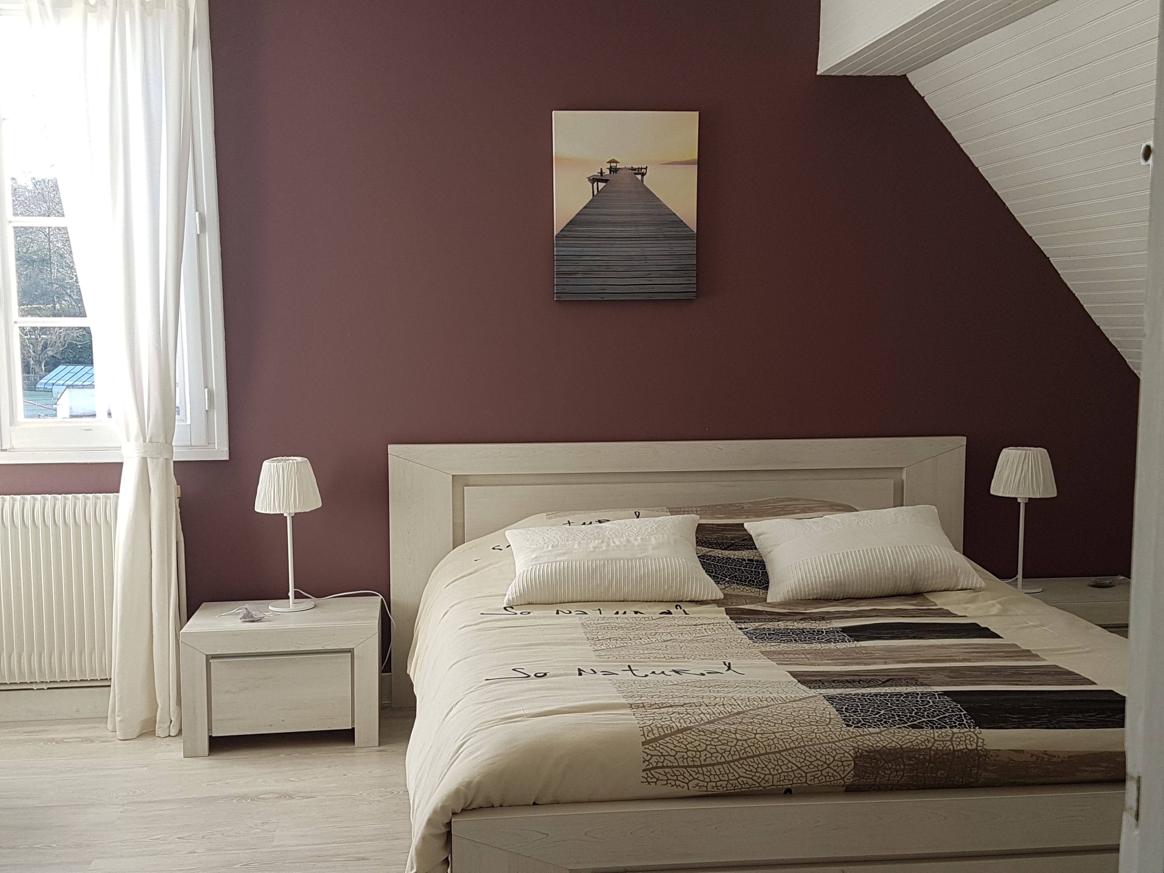 A bedroom with a bed, two bedside tables, a window with curtains, a painting on the wall, and a sloped ceiling.