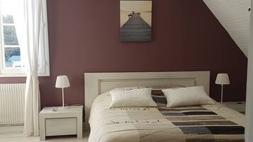 A bedroom with a bed, two bedside tables, a window with curtains, a painting on the wall, and a sloped ceiling.
