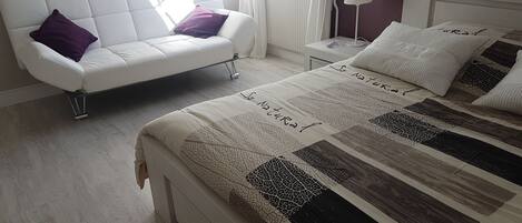 A bedroom with a bed, a white sofa, a nightstand, and a window with curtains.