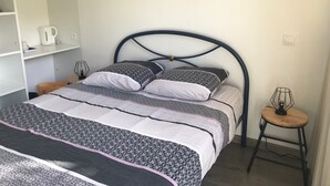 Ein ordentlich bezogenes Bett mit gemusterten Bettwäsche, ein Nachttisch mit Lampe und ein gerahmtes Bild mit Strandmotiv an der Wand.