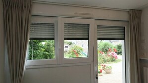 Eine Tür mit zwei Fenstern, jeweils mit Rollläden, die zu einem Garten mit Topfpflanzen und einem Weg führen.