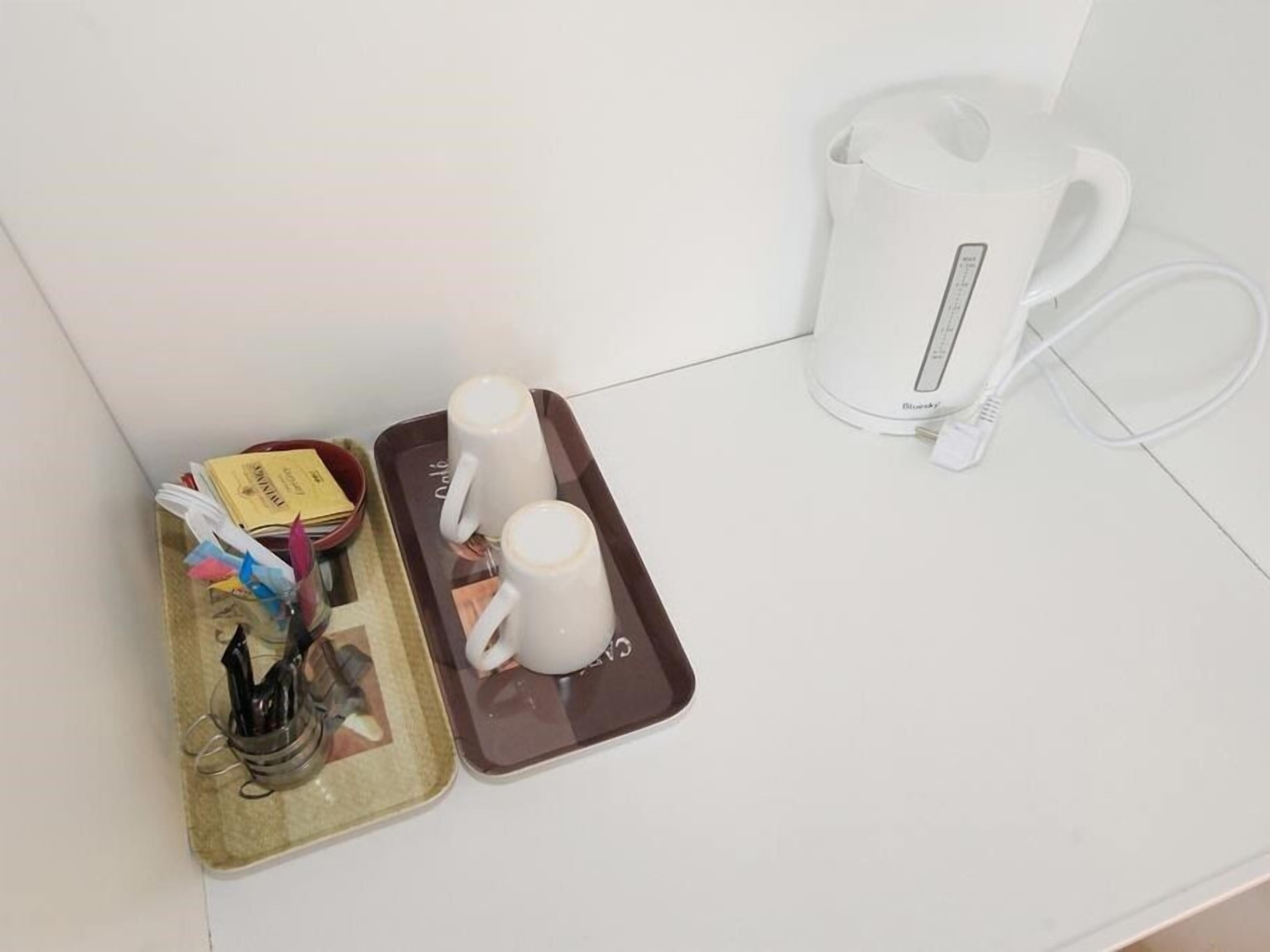 Una bandeja con dos tazas blancas, una tetera eléctrica blanca y un tazón pequeño con una cuchara sobre una encimera blanca.