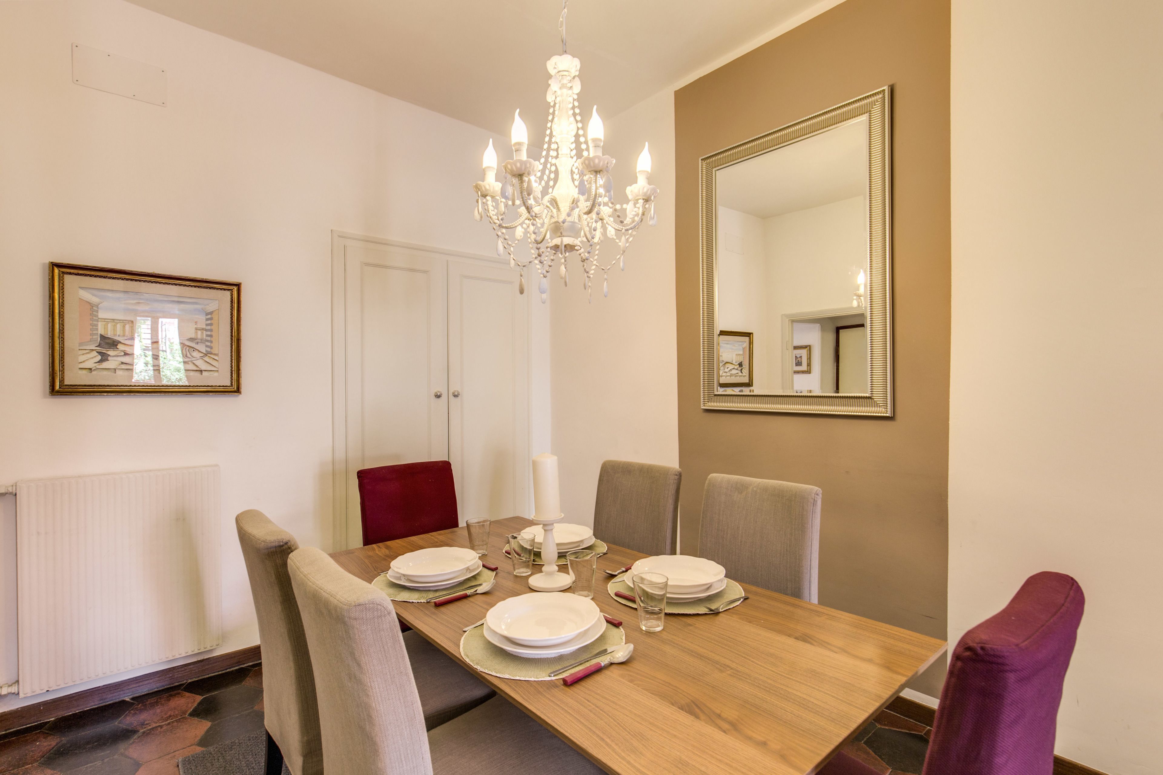 Une salle à manger avec une table en bois dressée pour quatre personnes, entourée de chaises. Un lustre est suspendu au plafond et un grand miroir orne le mur.