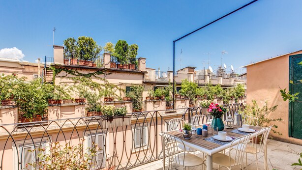 A rooftop terrace with a table set for dining, surrounded by potted plants and a view of neighboring buildings. - Spagna Glamour Life Penthouse (Rome)