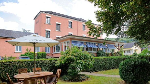 A hotel with a red facade, a covered outdoor seating area with a table and chairs, and a well-maintained garden. - Hôtel Gruber (Rosport-Mompach)