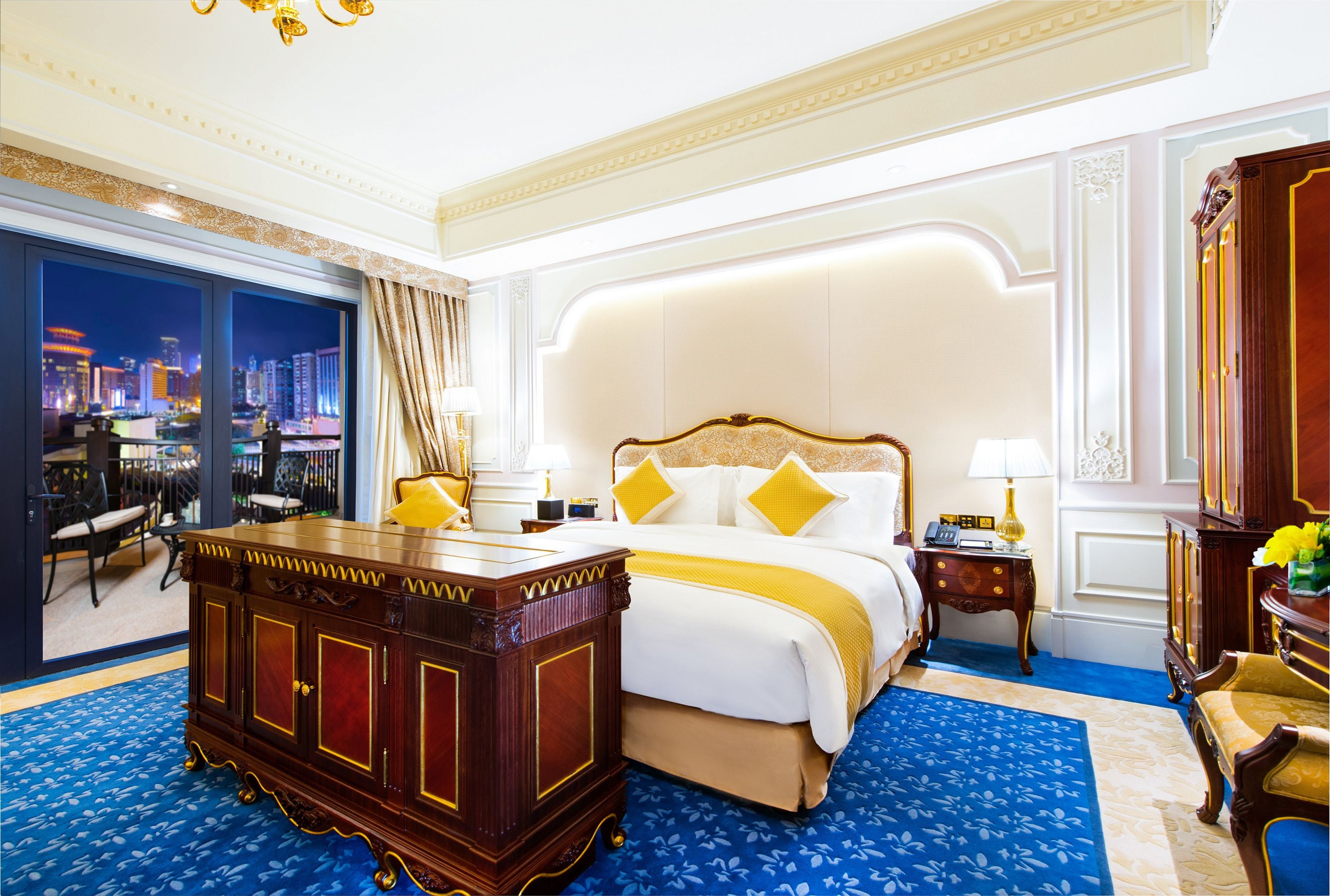A luxurious hotel room with a large bed, a wooden dresser, and a view of a cityscape at night.