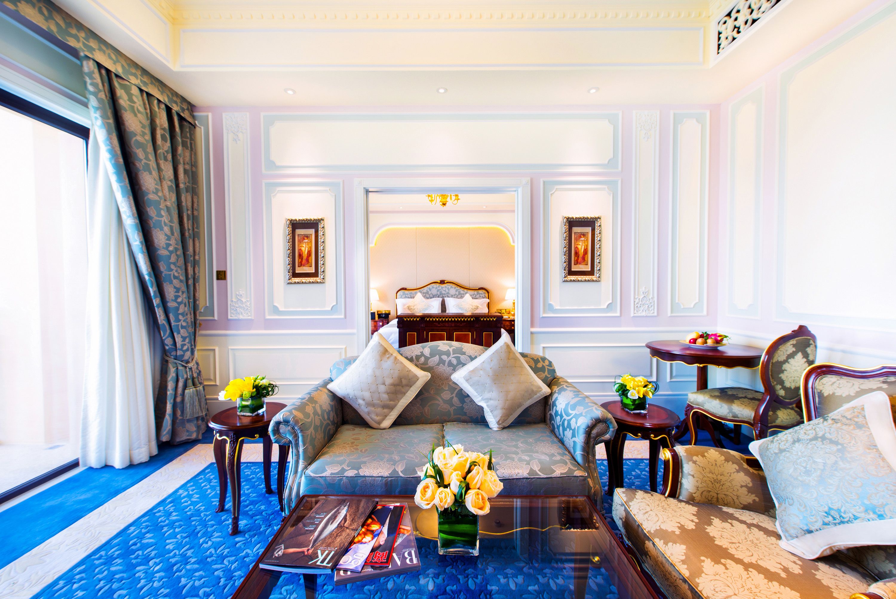 Een luxueuze woonkamer met een zithoek, een bijzettafeltje met bloemen, en uitzicht op een slaapkamer met een bed en een kroonluchter.