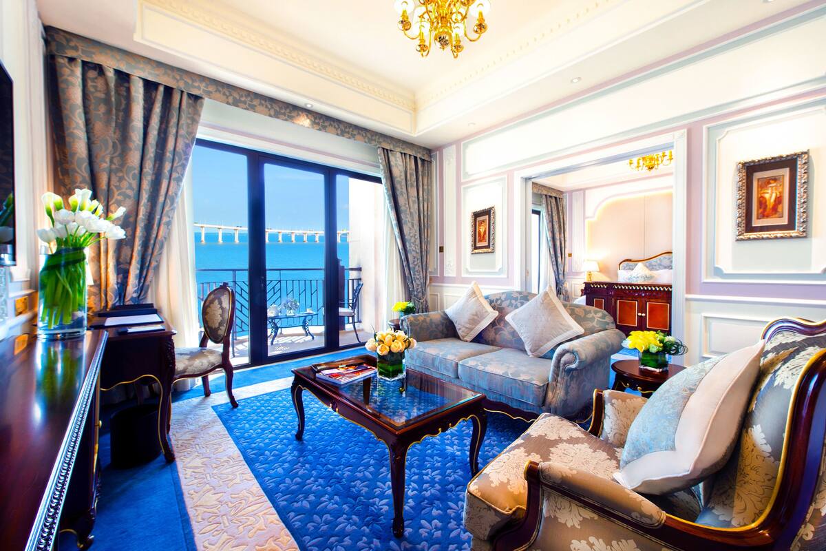 A spacious living room with a large window, a blue patterned rug, a wooden coffee table, and a sofa with patterned cushions.