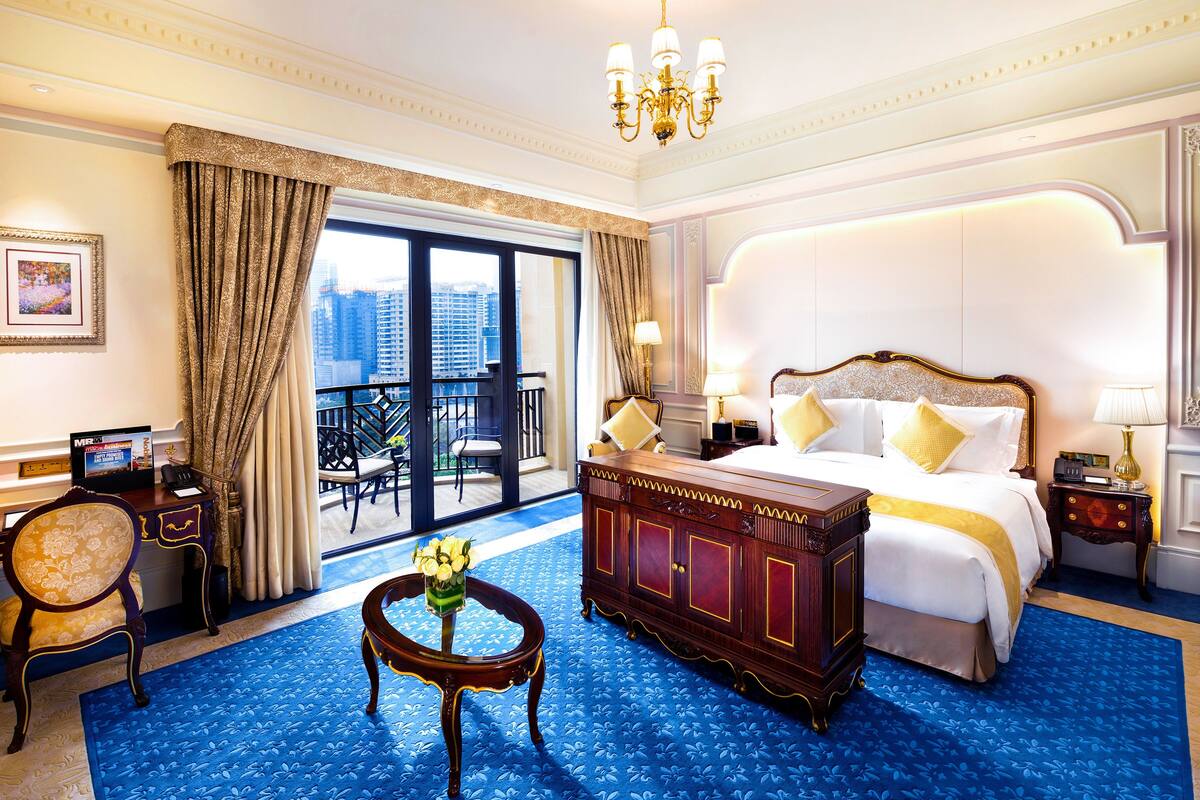A hotel room with a large bed, a desk, a chair, a small table with flowers, and a view of the city through the window.