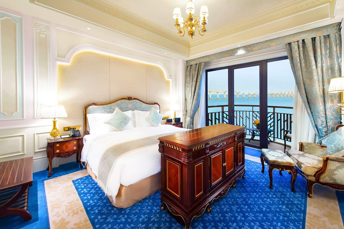 A luxurious hotel room with a large bed, a wooden dresser, a sitting area with a chair, and a balcony with a view of a bridge.