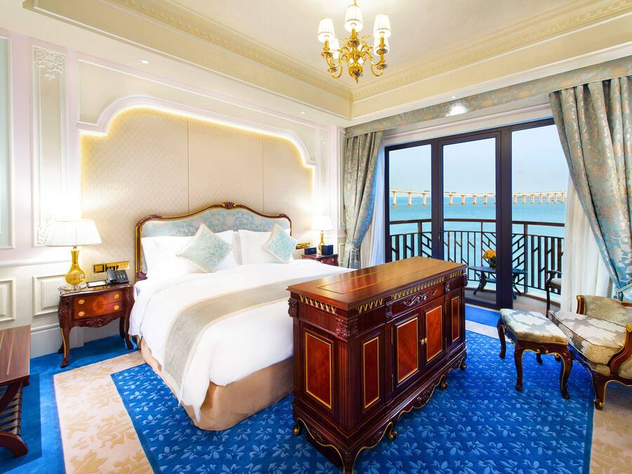 A luxurious hotel room with a large bed, a wooden dresser, a sitting area with a chair, and a balcony with a view of a bridge.