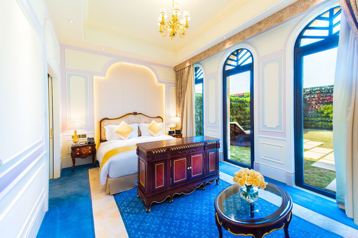 A luxurious bedroom with a large bed, a wooden dresser, a glass table with flowers, and a view of the garden through arched windows.