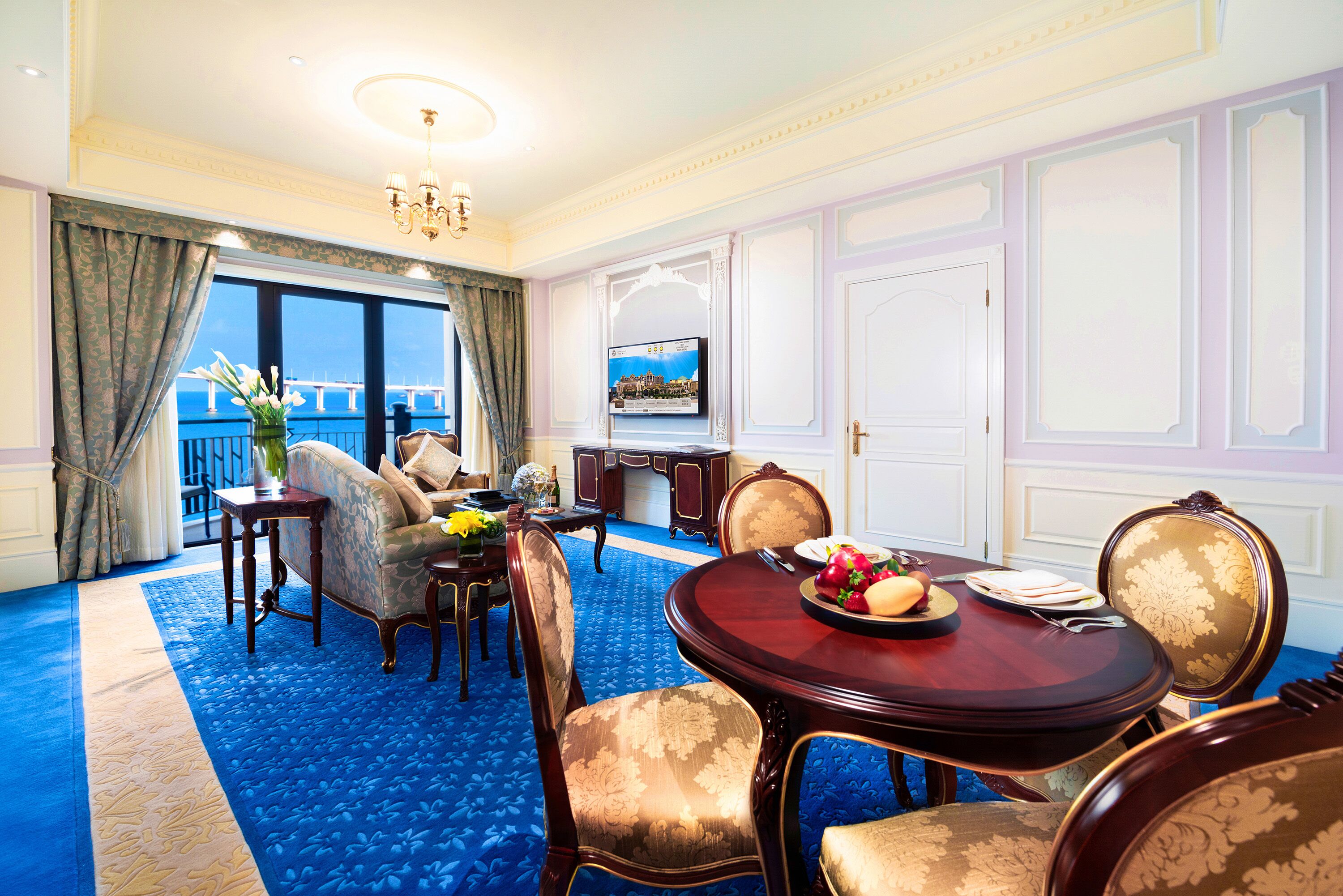Una habitación con alfombra azul, una mesa de comedor redonda con sillas, un sofá, un televisor y vistas al mar a través de amplias ventanas.