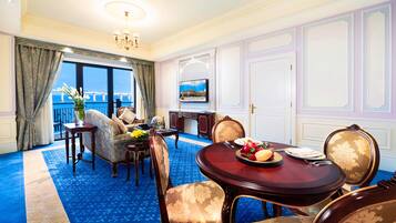 Una habitación con alfombra azul, una mesa de comedor redonda con sillas, un sofá, un televisor y vistas al mar a través de amplias ventanas.