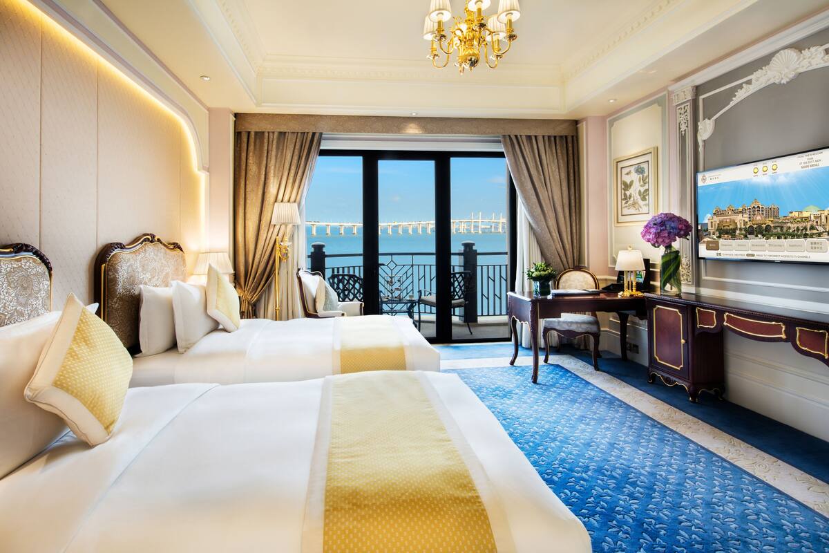 A hotel room with two beds, a desk, a chair, a TV, and a view of a bridge.