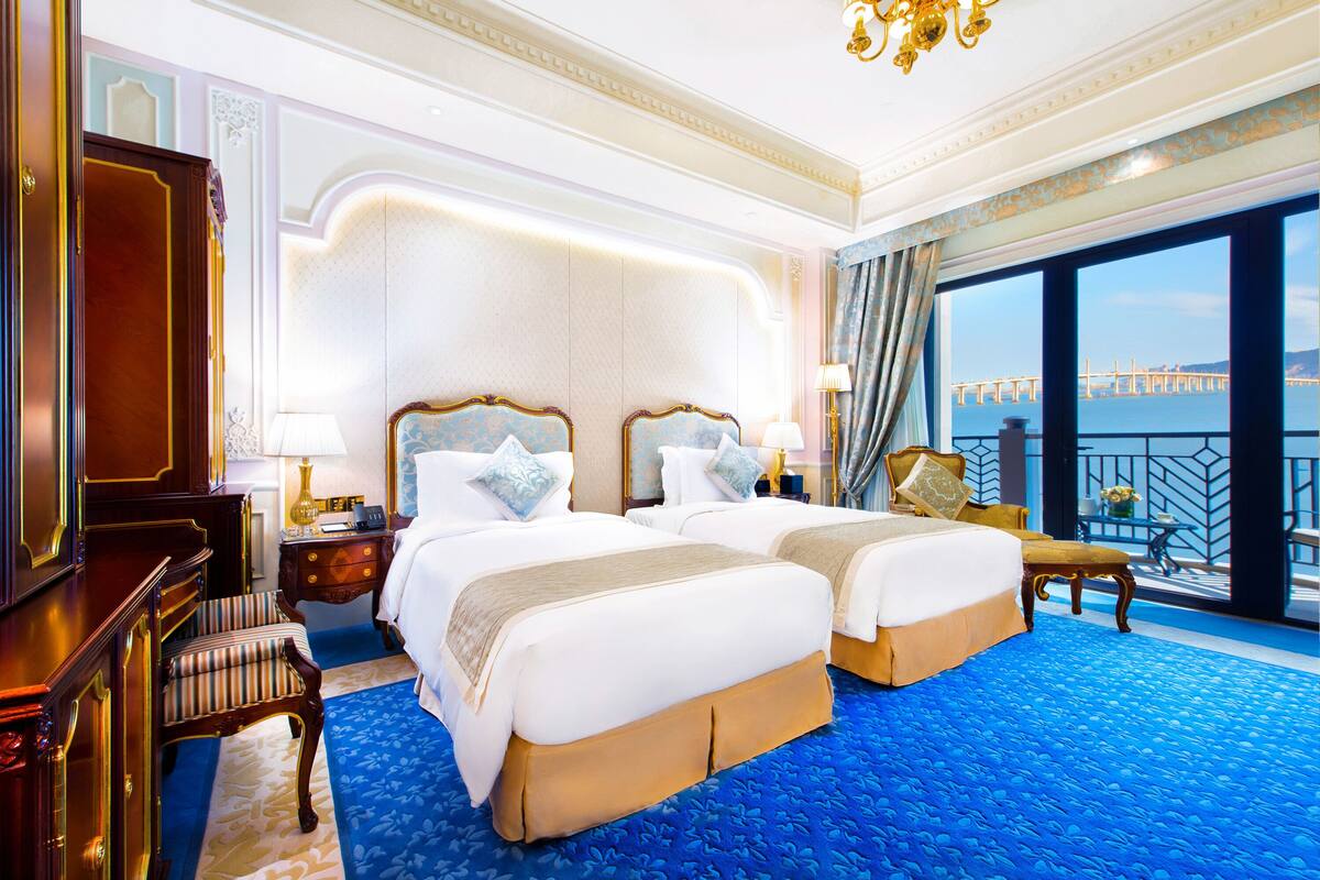 A luxurious hotel room with two large beds, a sitting area with a chair, a desk, and a view of a bridge.