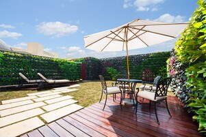 Un patio con una terraza de madera, una mesa redonda con sillas y un gran paraguas.