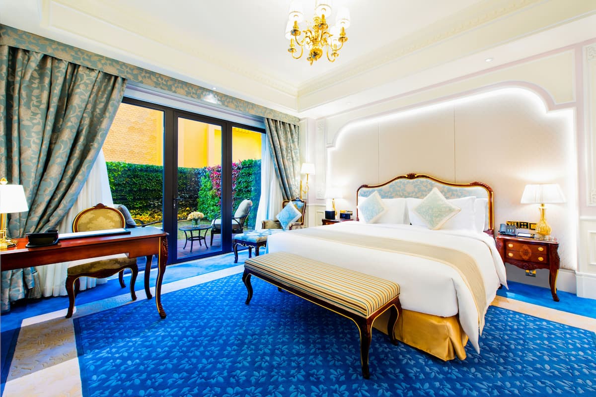 A spacious hotel room with a large bed, a desk, and a chair. There is a view of a garden through the window.