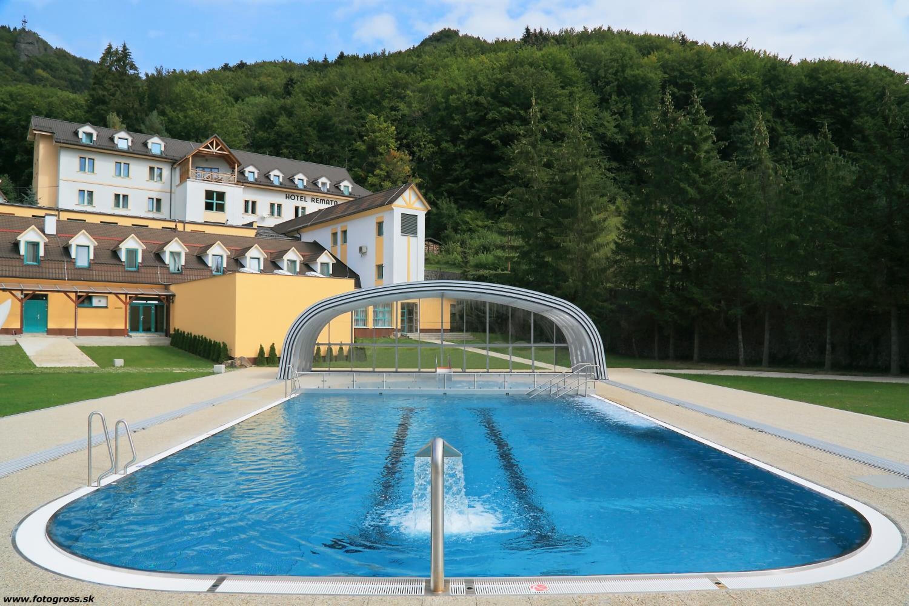A swimming pool with a diving board, surrounded by a hotel with multiple floors and a forested hillside in the background.