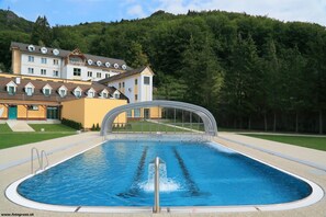 Una piscina con trampolín, rodeada por un hotel de varios pisos y un paisaje boscoso al fondo.