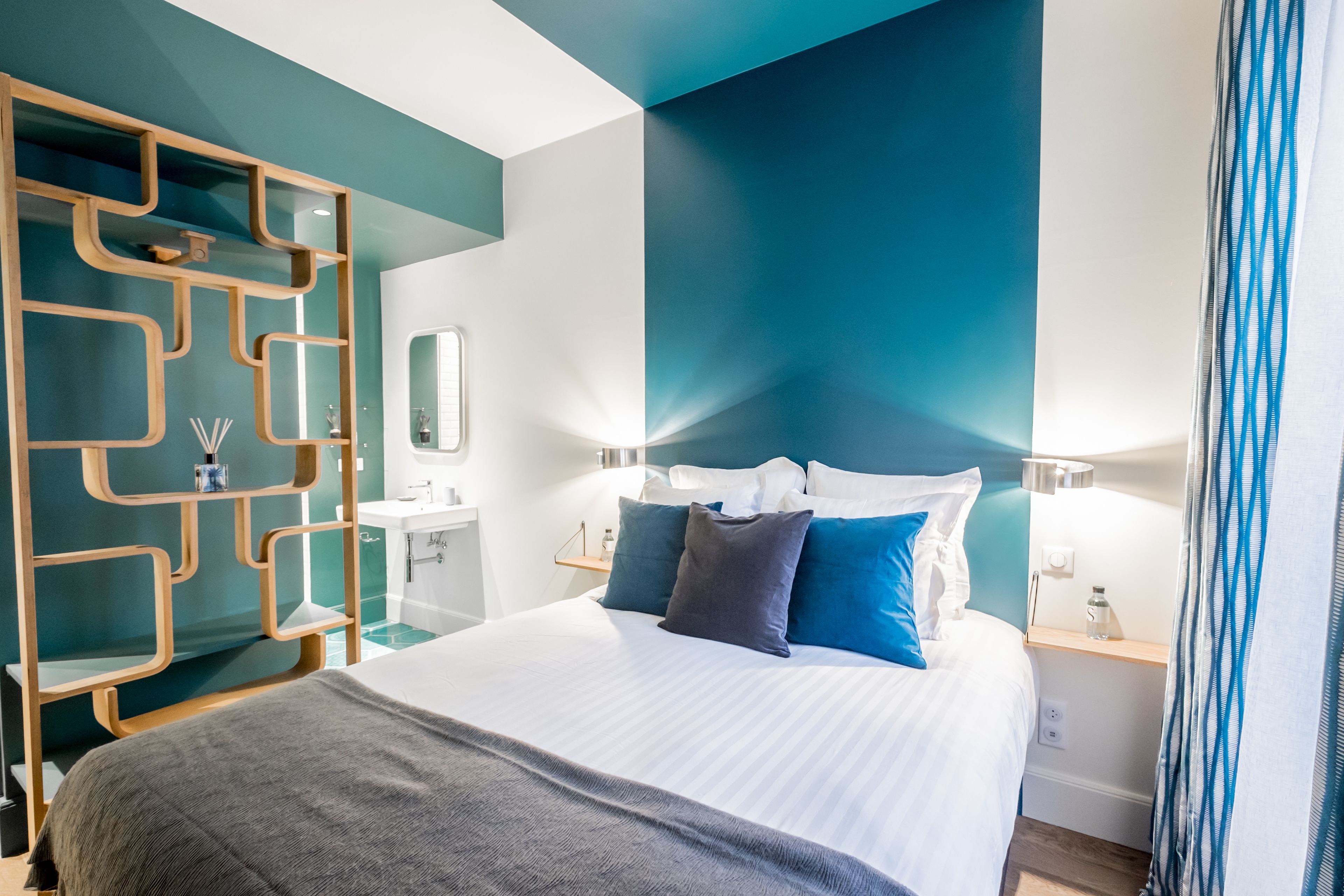A modern hotel room with a large bed, a wooden decorative screen, a bathroom with a sink and mirror, and a nightstand with a lamp.