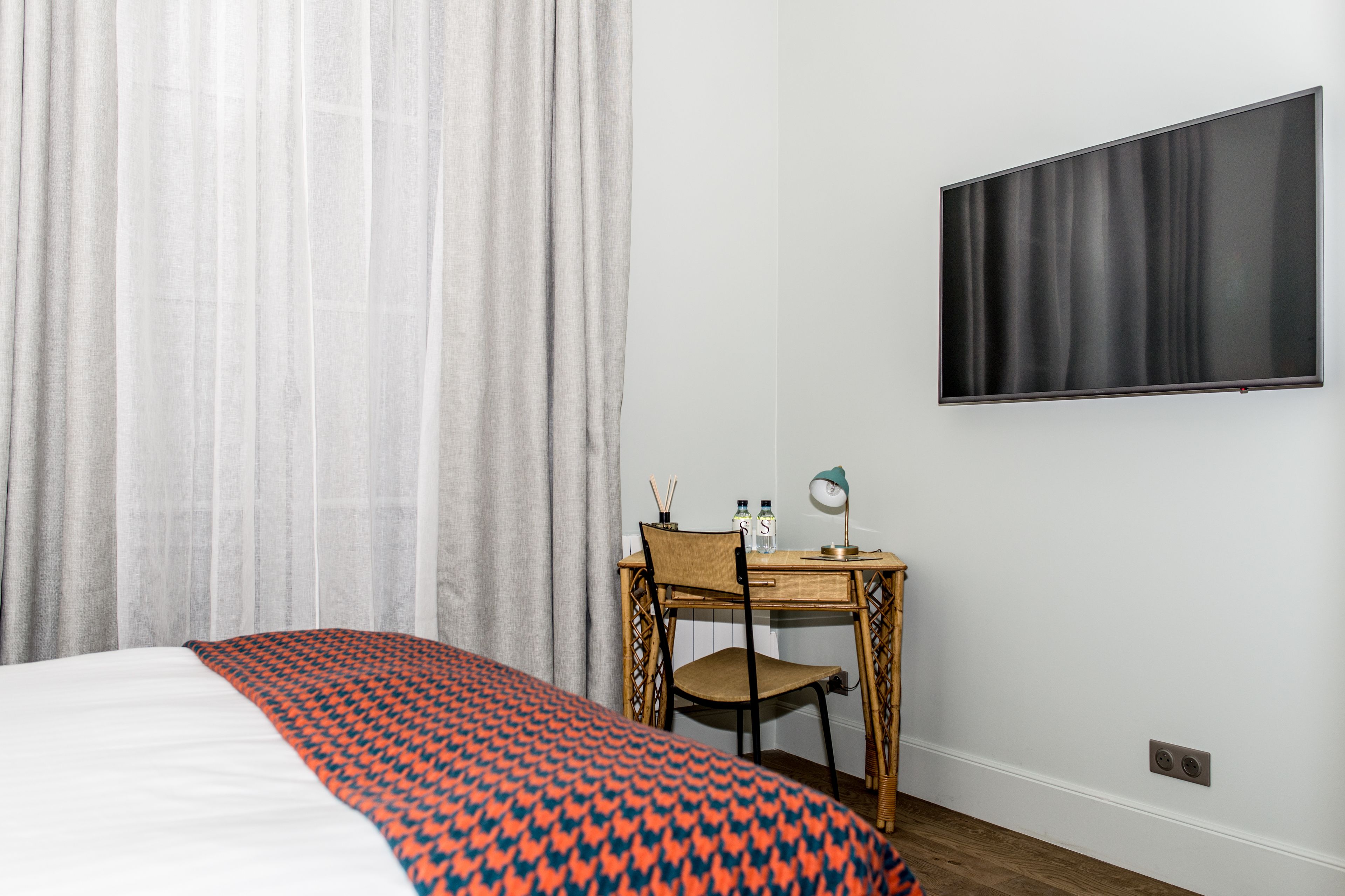 A hotel room with a bed, a desk with a chair, a flat-screen TV, and a window with curtains.
