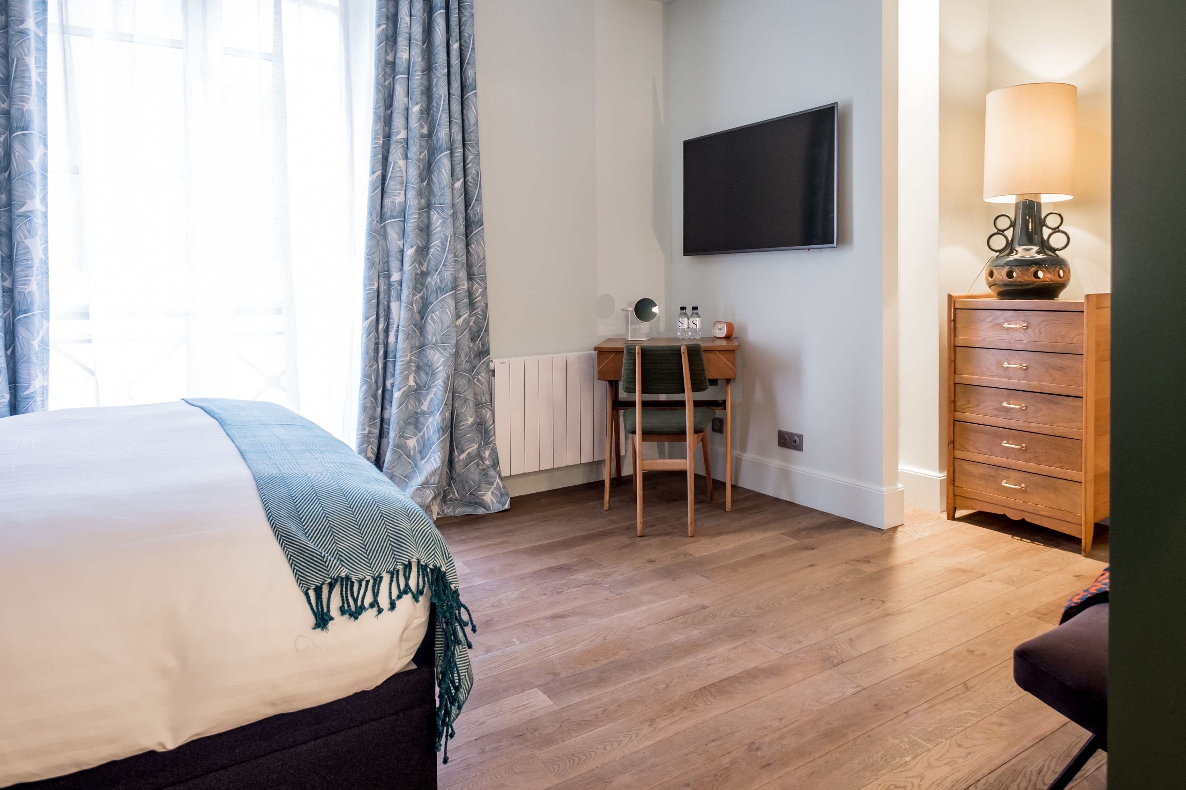 A hotel room with a bed, a desk, a chair, a TV, a lamp, and a dresser.