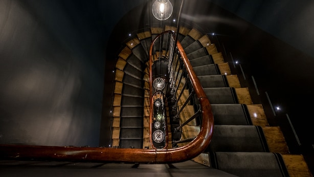 A spiral staircase with wooden steps and a central handrail, leading upwards in a dimly lit room.