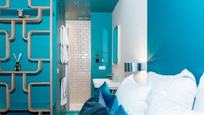 A modern hotel room with a teal color scheme, a large bed with white linens, a bathroom with a white sink and mirror, and a shower area.