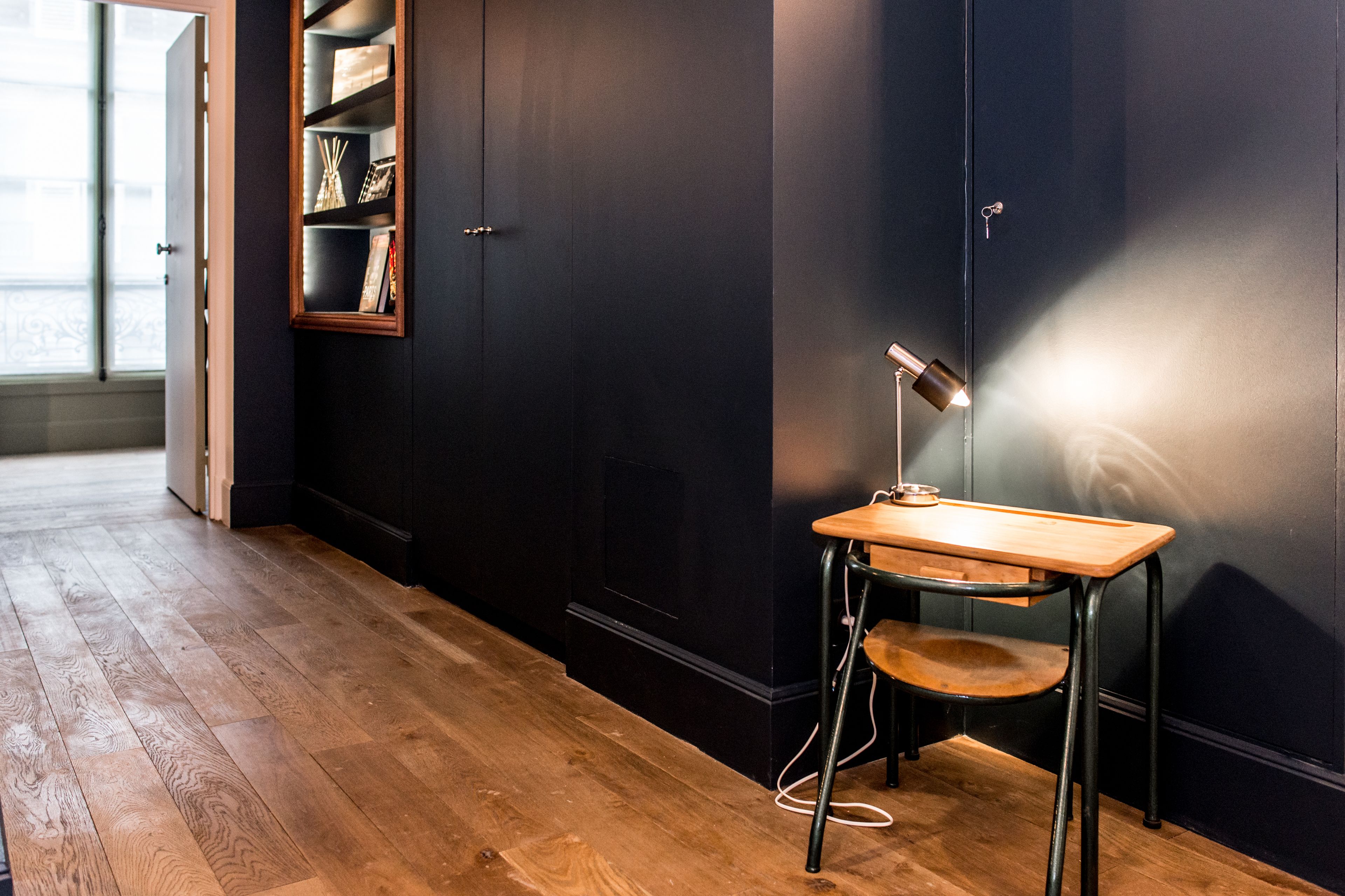 A room with a wooden floor, a desk with a lamp, and a bookshelf against a dark wall.