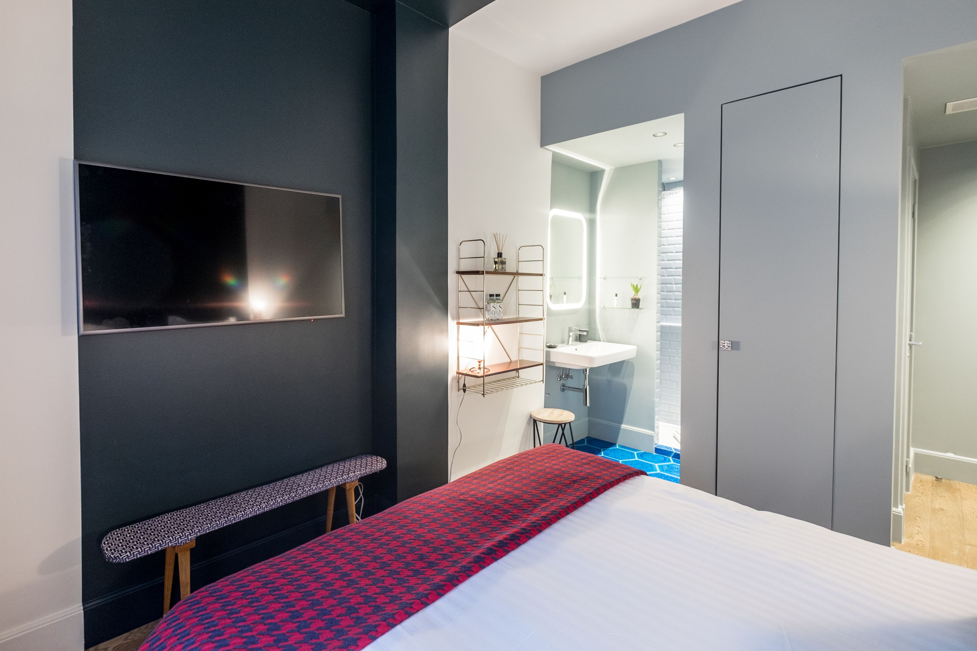 Habitación de hotel moderna con una cama grande, un banco, un televisor montado en la pared, un baño pequeño con espejo y lavamanos, y una puerta que da a otra habitación.