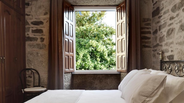 A bedroom with a bed, a chair, a window with wooden shutters, and stone walls.