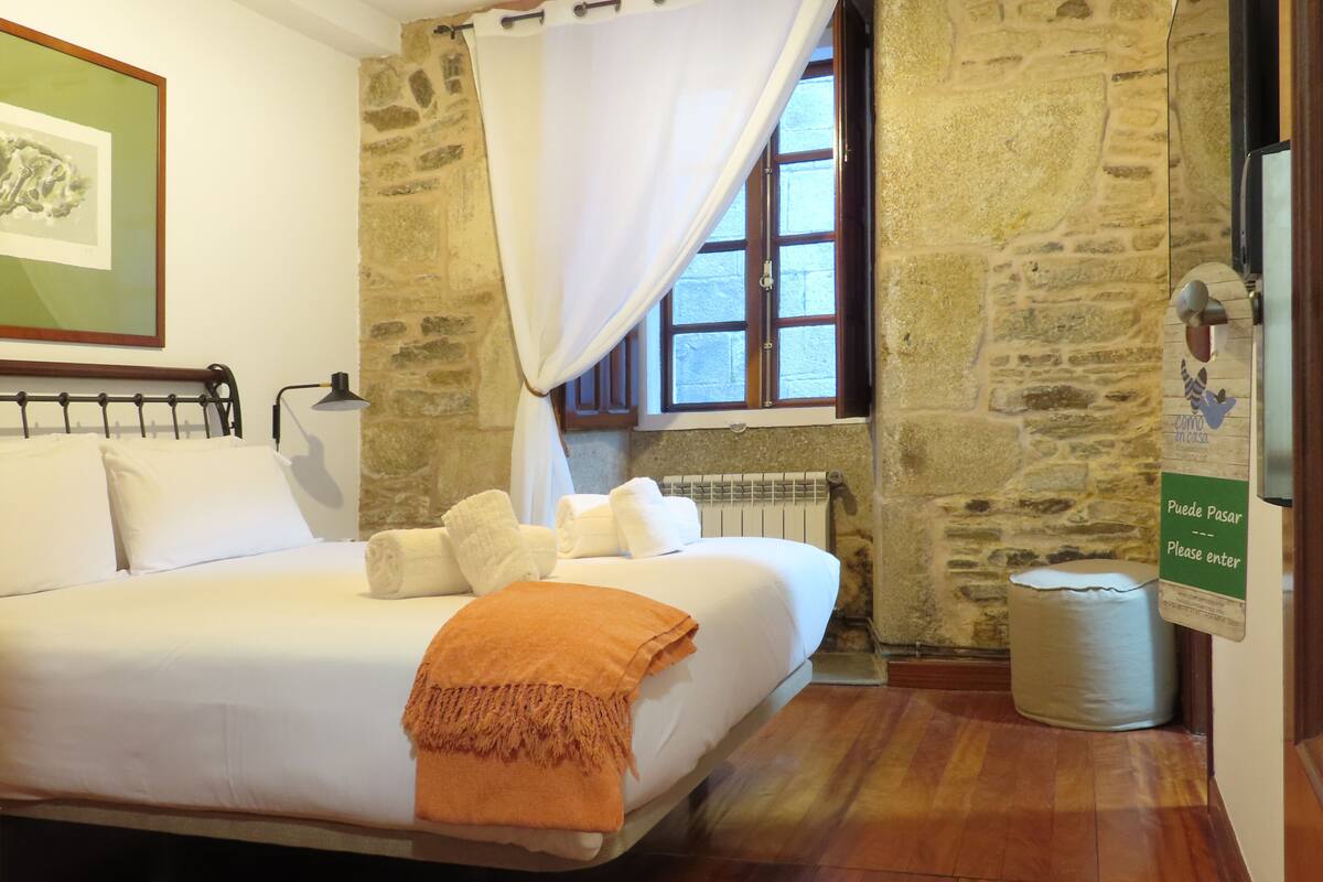 Una cama bien hecha con sábanas blancas y un plaid naranja, una pared de piedra, una ventana con cortina blanca y un cartel en la puerta.