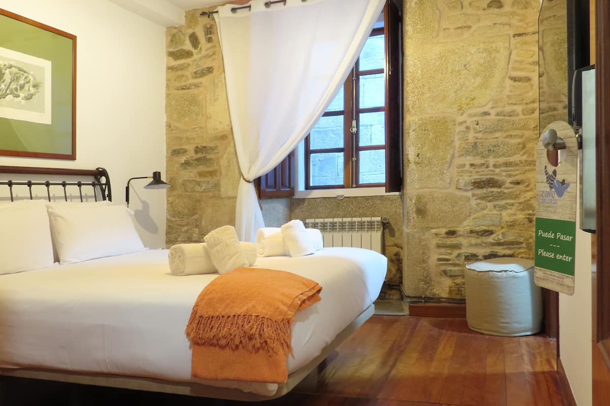 Un lit bien fait, avec du linge de lit blanc et un plaid orange, un mur en pierre, une fenêtre avec un rideau blanc et une pancarte sur la porte.