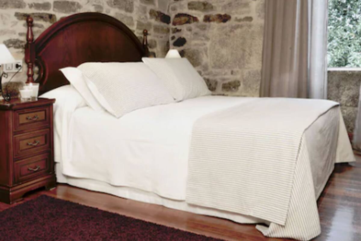 Una cama con sábanas blancas, cabecera de madera y alfombra rojo oscuro.