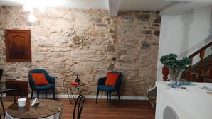 A room with a stone wall, two blue chairs with orange cushions, a round table with a white cup, a small black lamp, and a decorative sun-like wall piece.
