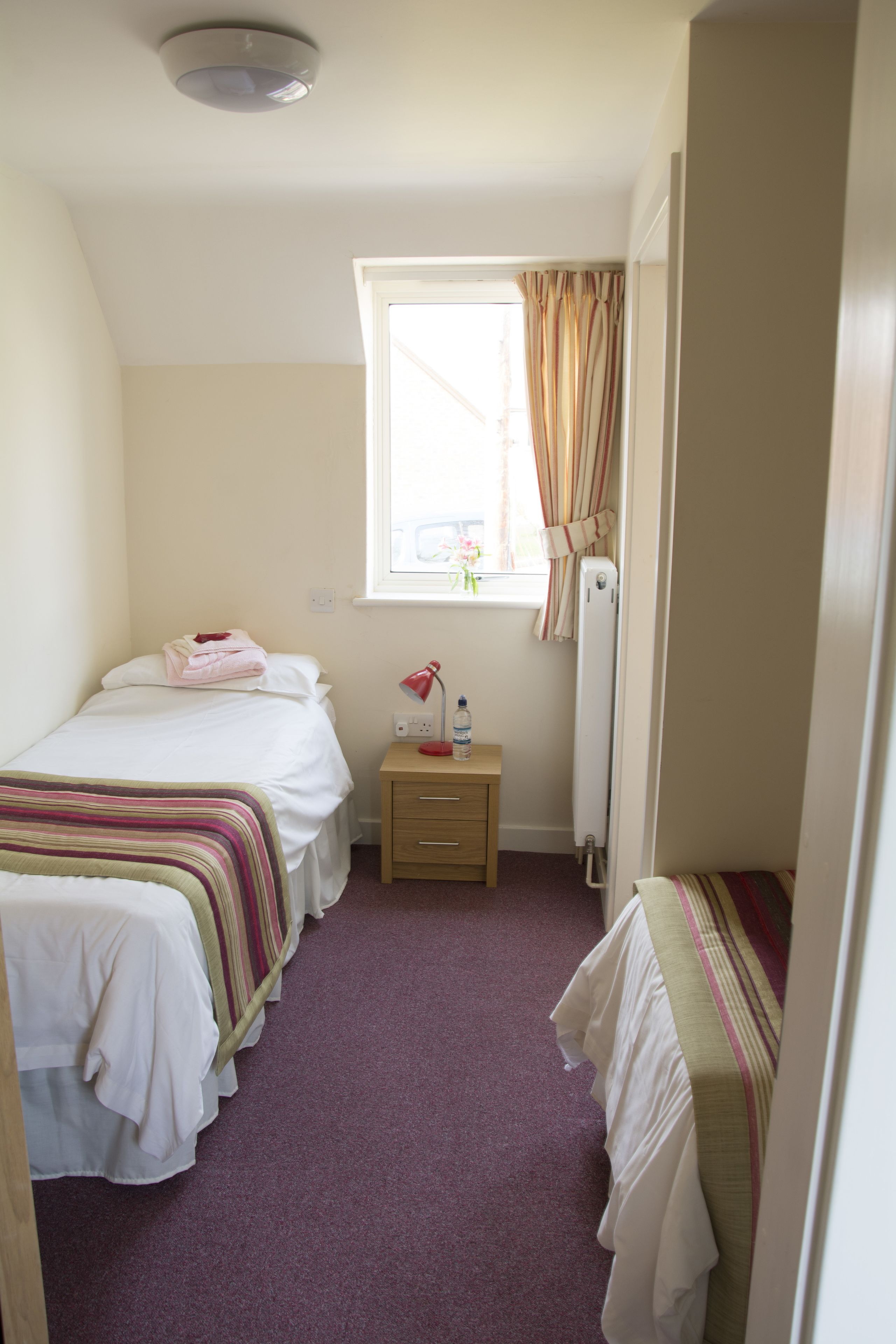 A small, well-lit bedroom with two beds, a bedside table with a lamp, and a window with curtains.