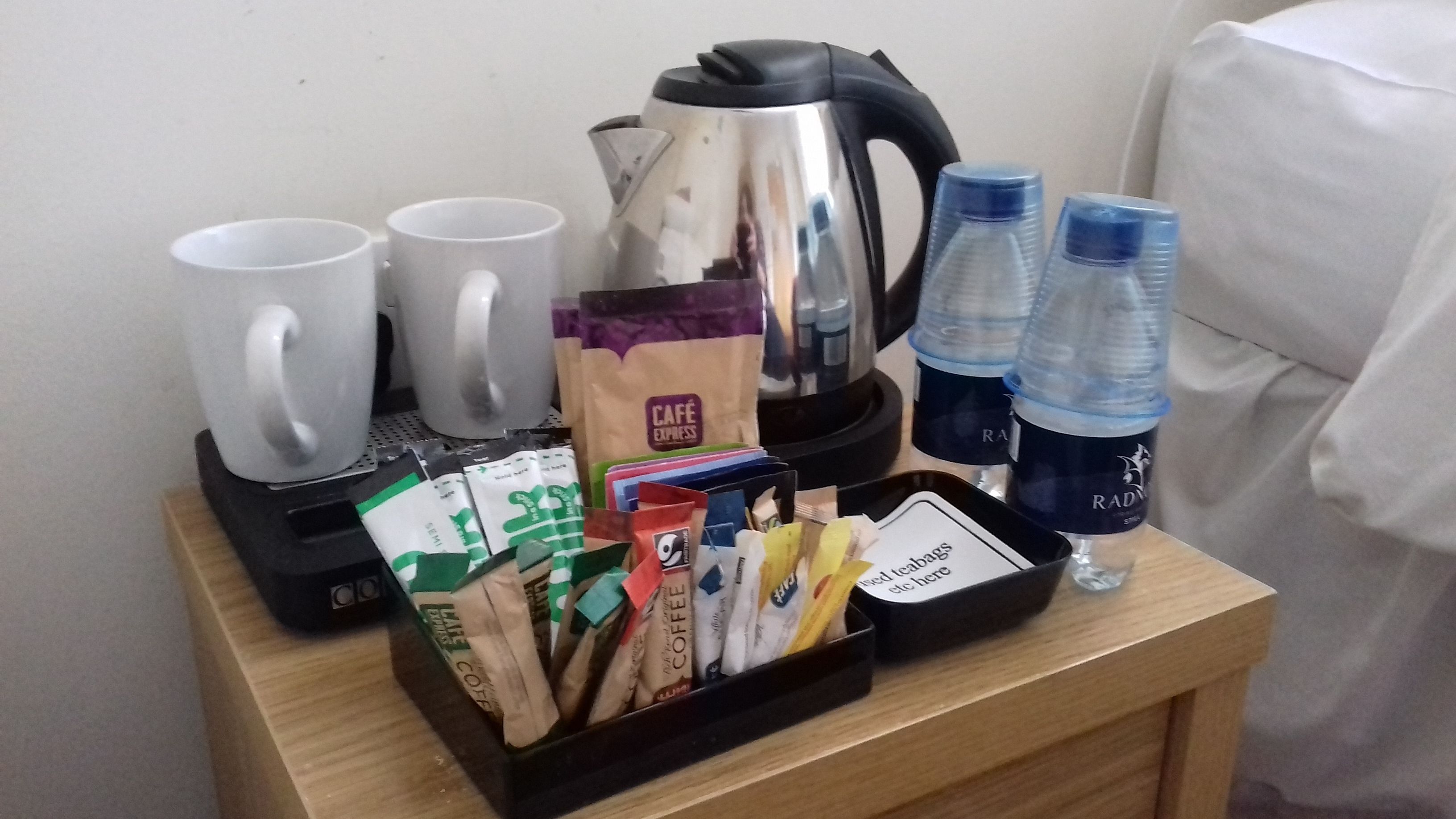 A breakfast setup with coffee packets, a kettle, mugs, and water bottles on a wooden table.