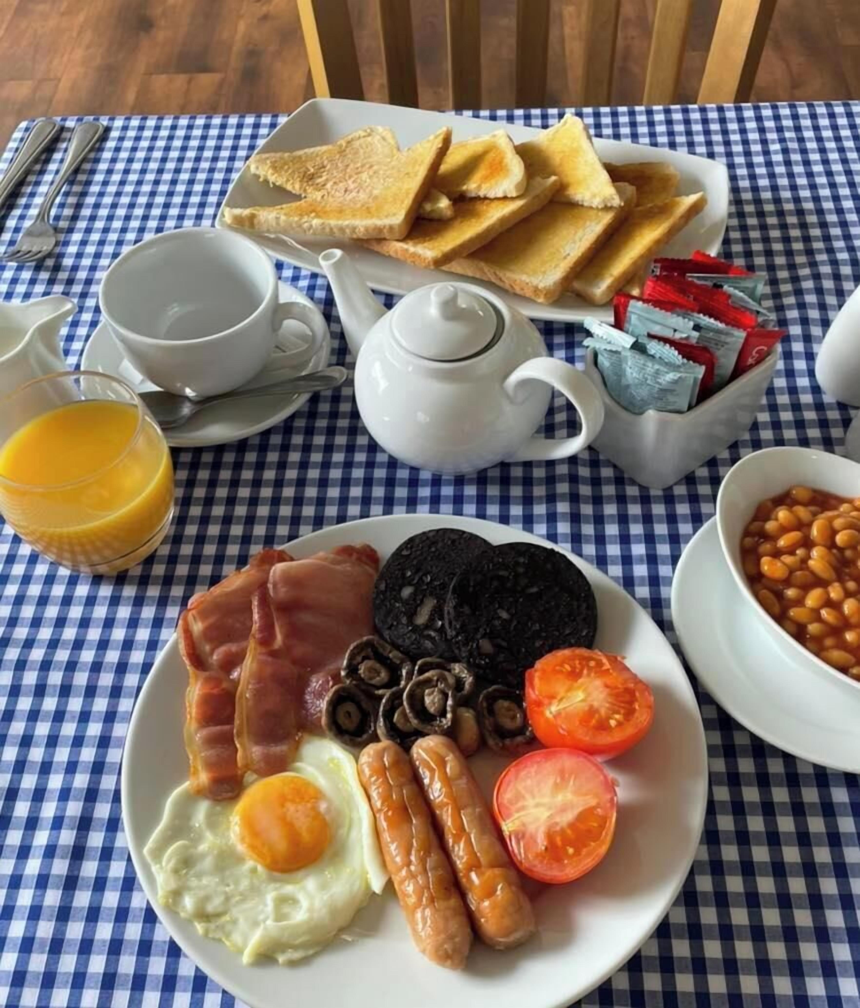 A breakfast plate with eggs, bacon, sausages, tomatoes, and beans, a teapot, a cup, and toast on a checkered tablecloth.