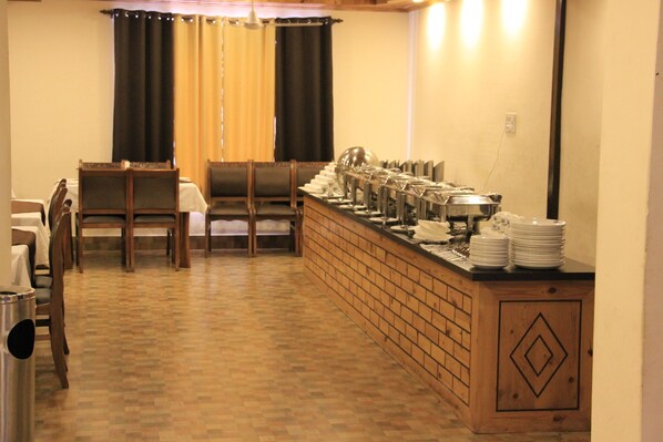 Disposição de buffet com um balcão comprido, várias cadeiras e uma mesa em um espaço com piso de madeira e parede de tijolos decorativa.