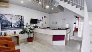 Zona de recepción de un hotel con suelo de mármol, mostrador de recepción, planta en maceta, televisor y escalera.